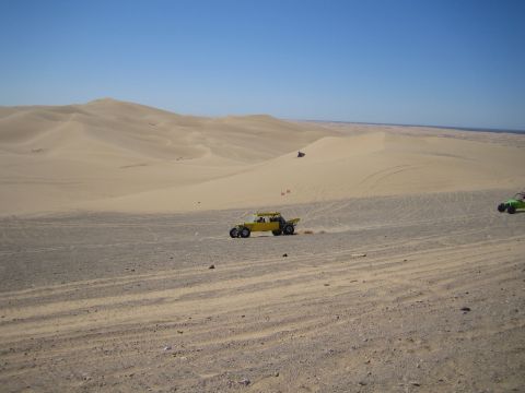 1968 Ford Mustang - Two dune buggies traverse a sandy desert with large dunes under a clear blue sky.