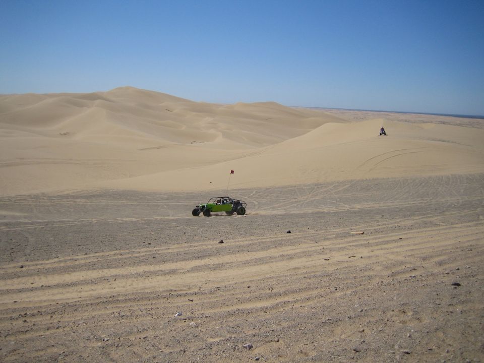 Green dune buggy with a red flag drives across a sandy desert landscape with large dunes under a clear blue sky