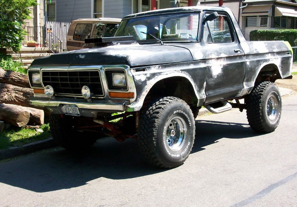 Lifted late-70s Ford pickup in primer with chrome wheels and mud tires parked on a residential street