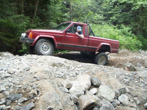 1985 Ford F-150 - A red 1986 Jeep Comanche pickup truck tackles a challenging rocky terrain on an off-road trail.