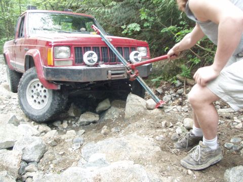 1985 Ford F-150 - A person uses a hi-lift jack to free a red Jeep Comanche pickup truck from a rocky obstacle on a trail.