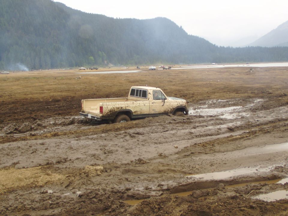 Beige pickup truck stuck in deep mud, front wheels submerged. Other vehicles visible in the distance