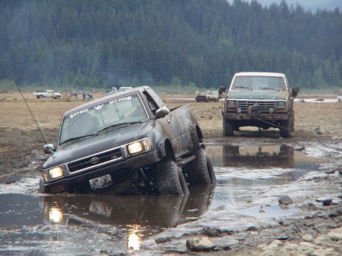 1985 Ford F-150 - Two trucks are stuck in a muddy area with trees in the background.