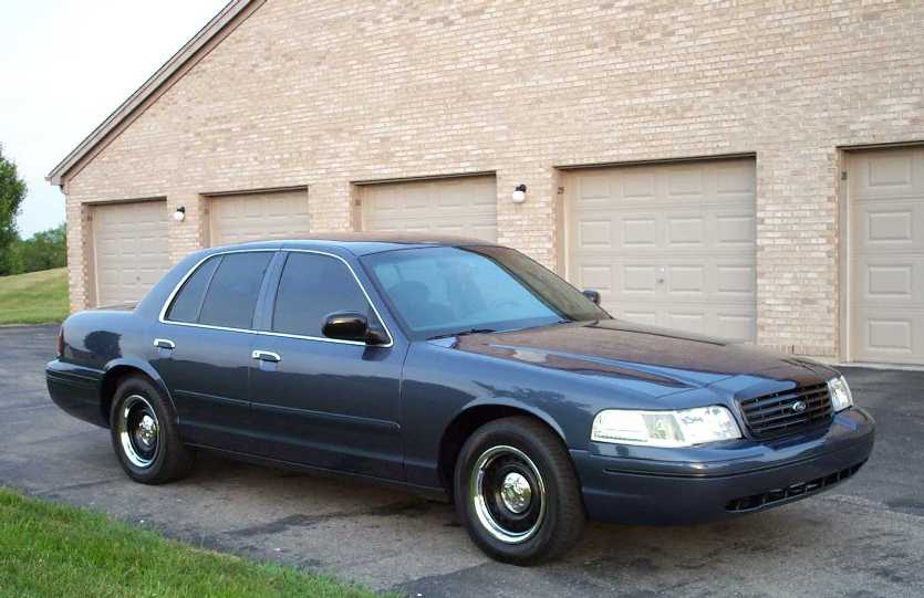 Dark blue-gray Ford Crown Victoria sedan, 3/4 front view, parked before a brick garage building