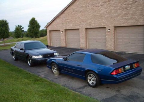 1998 Ford Crown Victoria - A dark blue 1998 Ford Crown Victoria is parked next to a blue coupe in front of a brick garage.