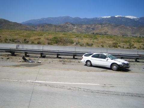 1990 Ford Bronco - white station wagon with heavy front-end crash damage rests on a concrete freeway shoulder against a guardrail, mountains behind.