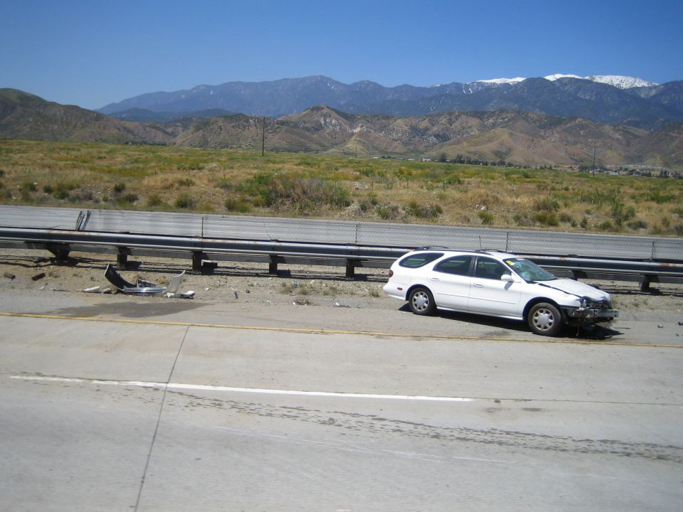 White station wagon with crushed front end stopped against a guardrail on a freeway shoulder