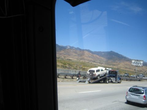 1990 Ford Bronco - Through a tinted window, a flatbed tow truck carries a damaged white SUV along a freeway beside dry mountains.