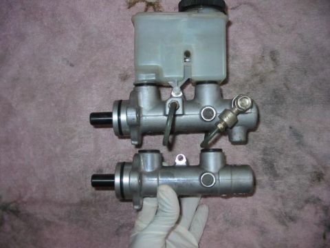 1995 Ford Probe - A gloved hand holds a brake master cylinder next to another one with a fluid reservoir.