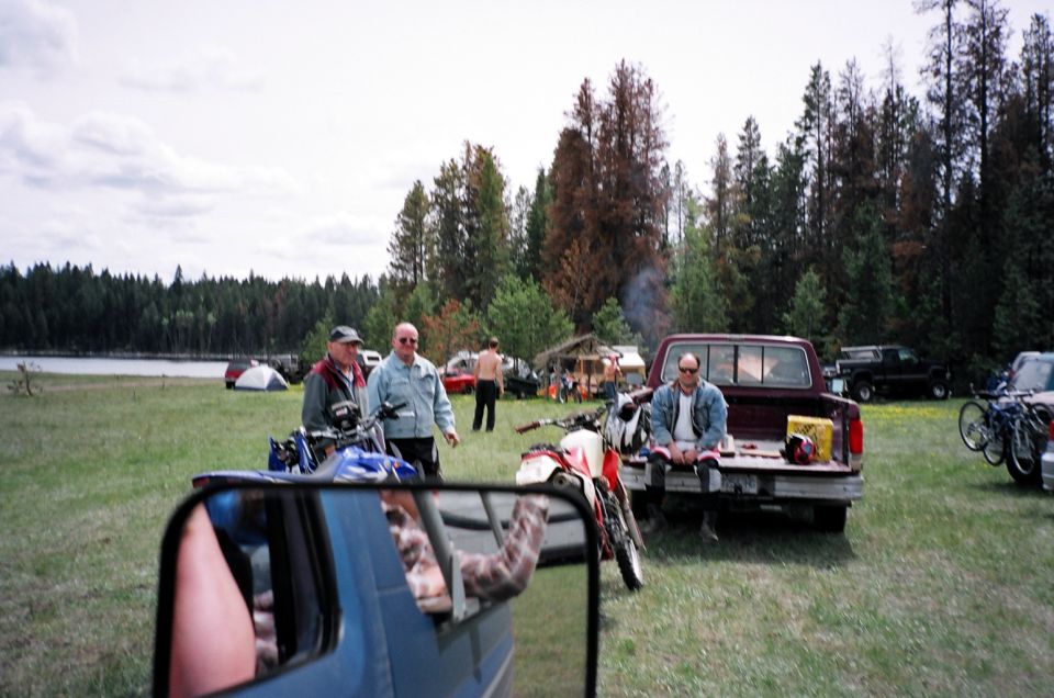 Men by dirt bikes and a red pickup with a person in the bed, lakeside meadow camp, mirror in foreground