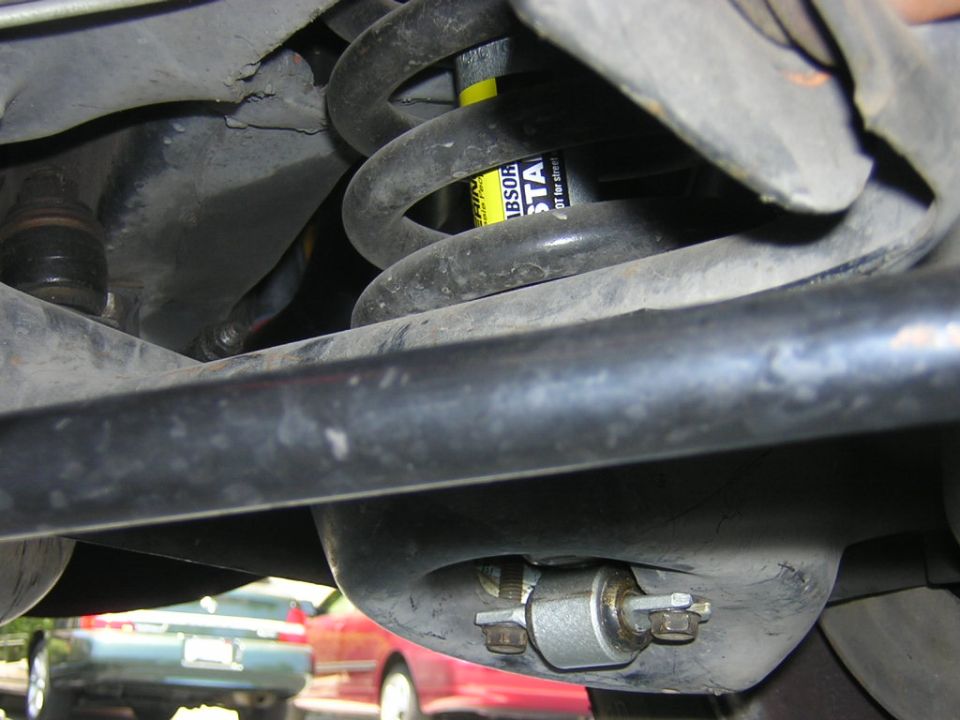 Car suspension with yellow shock absorber and black coil spring. Two cars visible in background