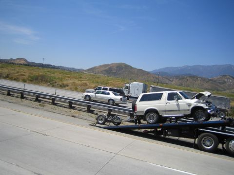 1990 Ford Bronco - white full-size SUV with front damage rides on a flatbed trailer down a freeway, with other vehicles and mountains beyond.
