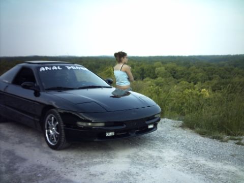 1995 Ford Probe - A black Ford Probe is parked on a dirt road with a woman sitting on its hood, overlooking a dense forest.