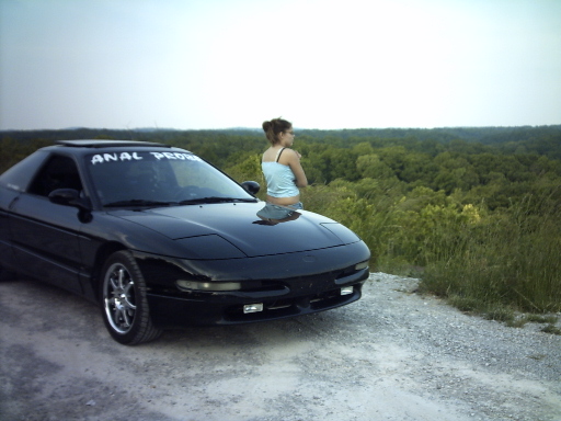 Black Ford Probe parked on a dirt road with a woman on the hood, forest in the background