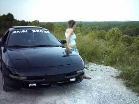 1995 Ford Probe - A black 1995 Ford Probe is parked on a gravel road with a woman leaning against it, overlooking a wooded landscape.