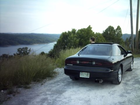 1995 Ford Probe - A black 1995 Ford Probe is parked on a dirt road with a scenic view of a lake and surrounding hills.
