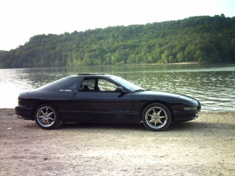 1995 Ford Probe - A black 1995 Ford Probe is parked on the shore of a lake with a wooded hillside in the background.