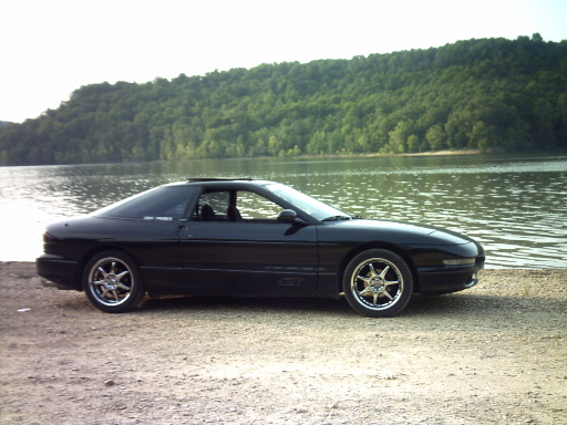 Black 1995 Ford Probe parked on a gravel shore next to a lake