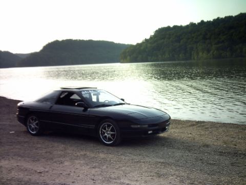 1995 Ford Probe - A black 1995 Ford Probe sits parked on a gravelly shore next to a calm lake, with a tree-covered hill in the background.