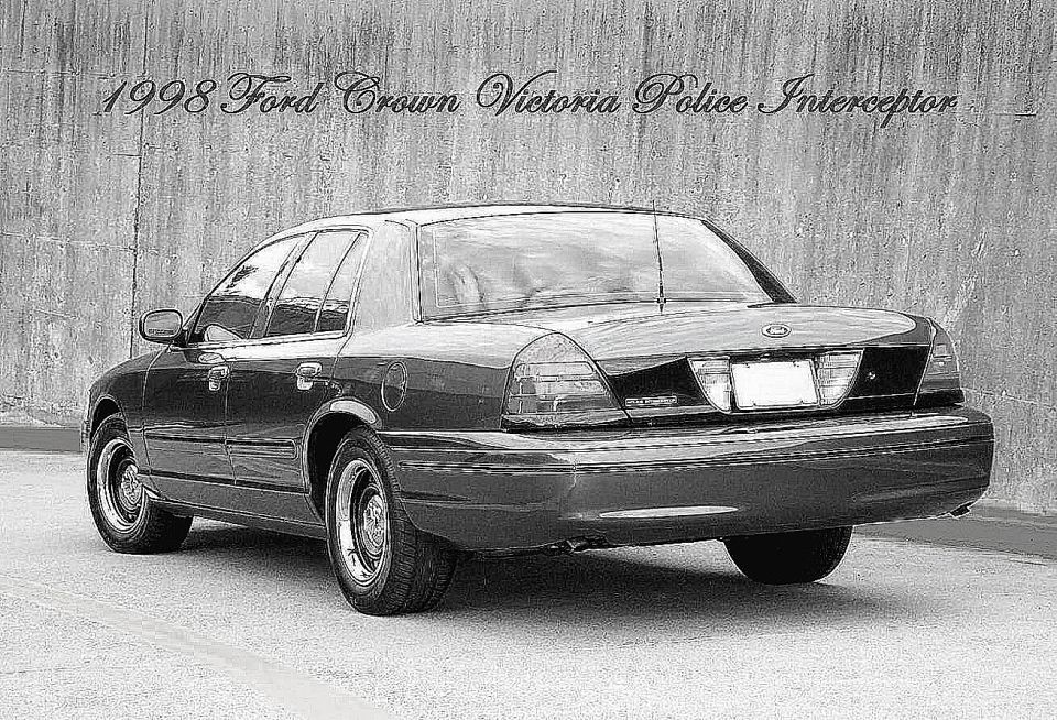 Rear three-quarter view of a dark 1998 Ford Crown Victoria sedan on steel wheels beside a concrete wall, black-and-white photo.