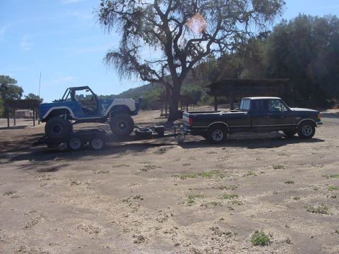 1971 Ford Bronco - A blue and white off-road vehicle is on a trailer, being towed by a black pickup truck under a large tree.