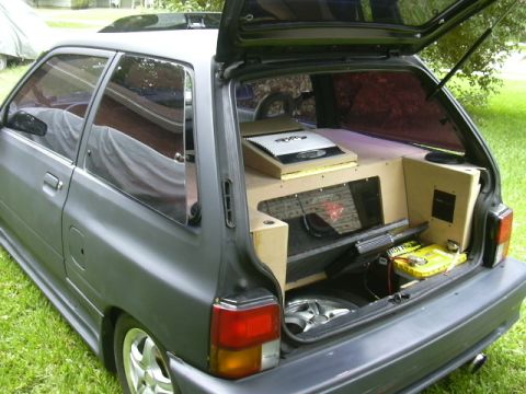 1991 Ford Festiva - The rear of a gray hatchback is open, revealing a custom sound system with multiple speakers and amplifiers installed in the cargo area.