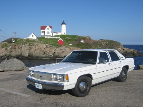 1991 Ford LTD Crown Victoria - Front three-quarter view of a white Ford LTD Crown Victoria sedan on black steel wheels, parked on a paved coastal overlook in front of the…
