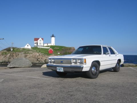 1991 Ford LTD Crown Victoria - White Ford Crown Victoria sedan parked in a coastal lot, shot from the 3/4 front left, with the Nubble lighthouse and its red-roofed keeper…