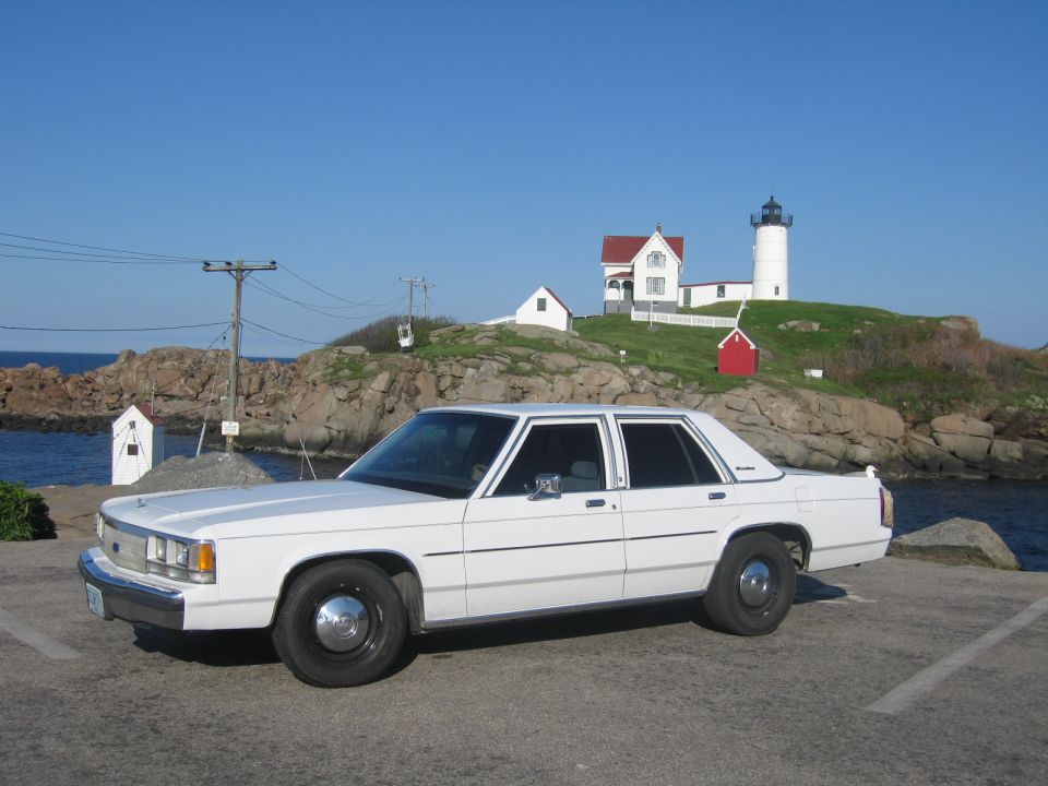 White sedan parked on asphalt with a lighthouse and ocean in the background