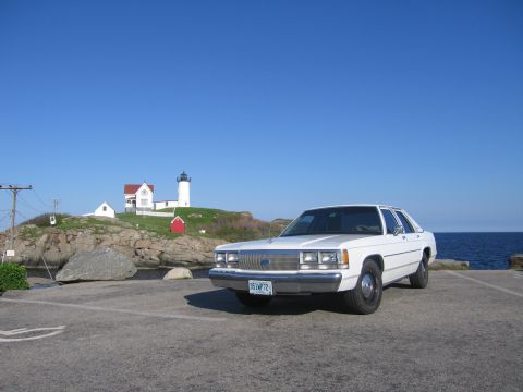 1991 Ford LTD Crown Victoria - A white Ford Crown Victoria four-door sedan parked at a coastal overlook under a clear blue sky, with a white lighthouse, red-roofed keeper…
