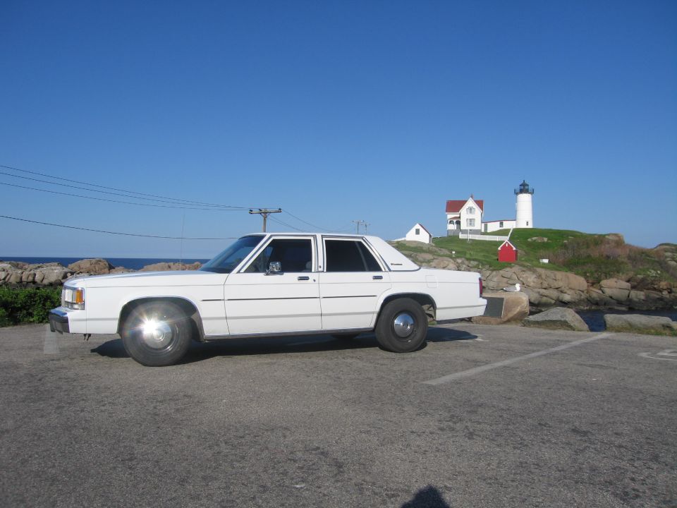 White sedan parked on asphalt with a lighthouse and ocean in the background