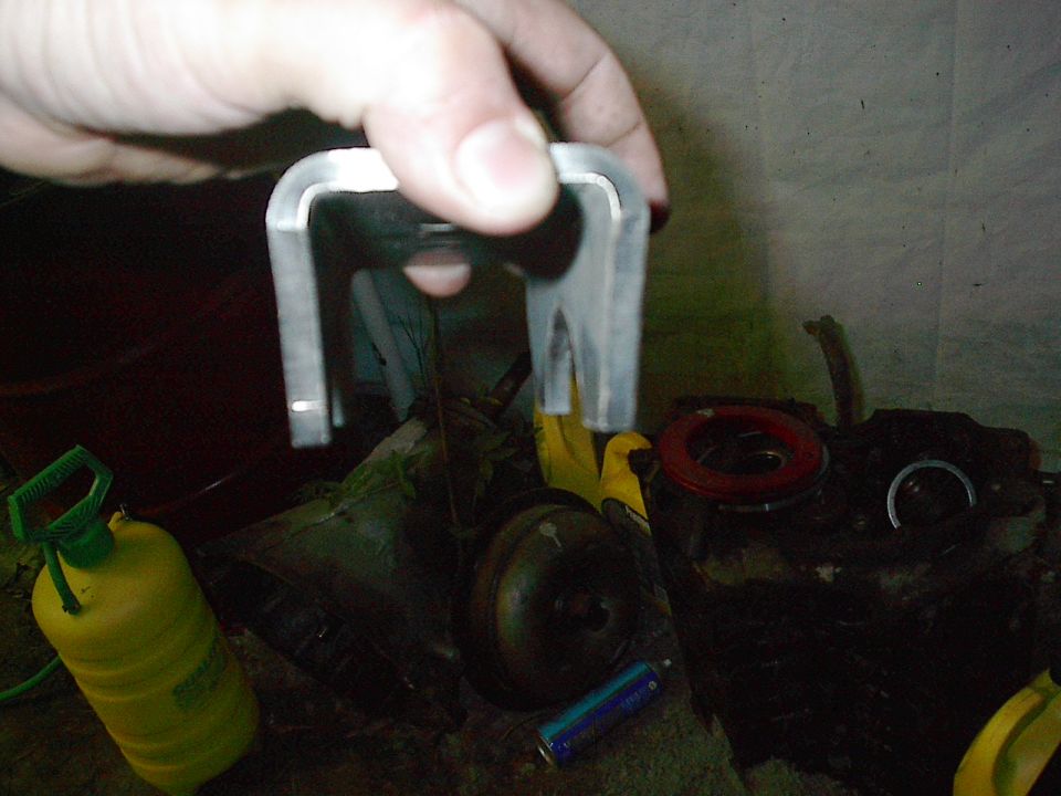 Hand holding a metal bracket over some vehicle parts, including a yellow spray bottle and a red ring