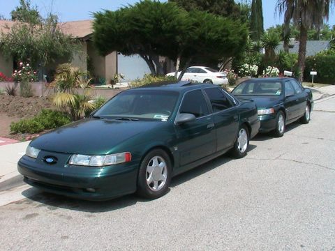 1993 Ford Taurus - Two green sedans are parked on a residential street. The front car is a 1996 Ford Taurus.