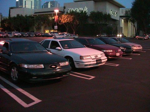 1993 Ford Taurus - A row of Ford Taurus sedans, including a 1993 model, parked outside a shopping mall at dusk.