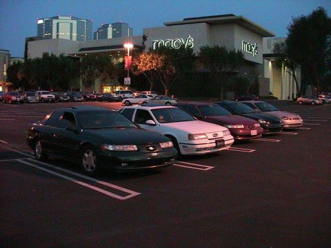 1993 Ford Taurus - A row of cars are parked in a lot outside a Macy's store at dusk.