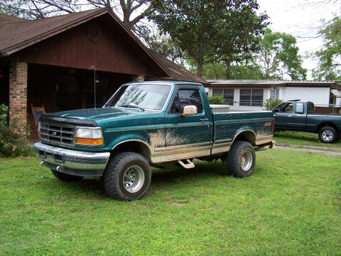 1996 Ford F-150 - A green pickup truck is parked on a grassy lawn next to a house, with another truck in the background.