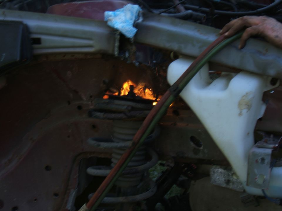 Car engine bay on fire, with a hand holding a hose and a white fluid reservoir visible
