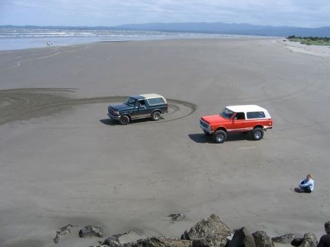 1979 Ford Bronco - Two SUVs drive on a beach with the ocean and distant hills visible.