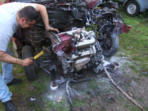 1998 Ford Taurus - A man uses a hammer on the engine of a heavily damaged vehicle, with another car partially visible in the background.