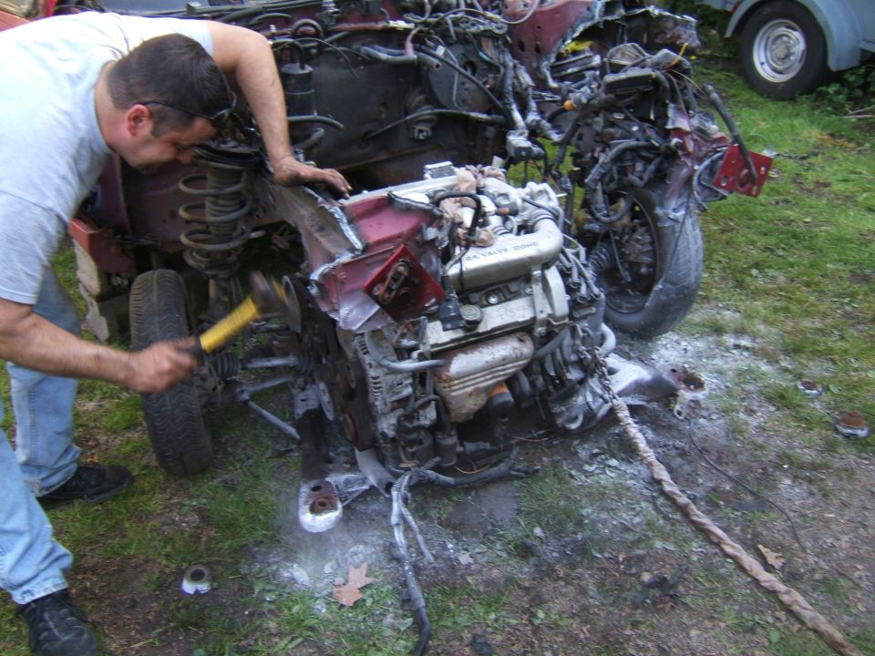 Man with hammer working on wrecked car engine. Another vehicle visible in background