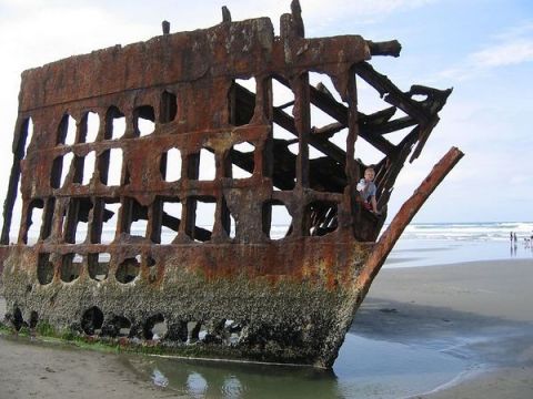 1979 Ford Bronco - A child sits within the decaying remains of a shipwreck resting on a sandy beach, with the ocean waves in the background.