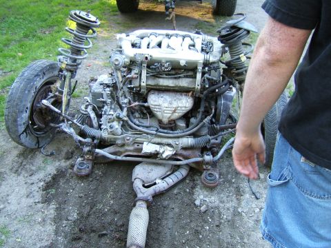 1998 Ford Taurus - Disassembled car engine and front suspension assembly lies on the ground, with a person observing.
