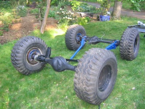 1978 Ford Bronco - Two bare solid axles sitting on a grassy lawn, each still wearing mud-terrain bias tires on black steel wheels. The front axle carries blue…