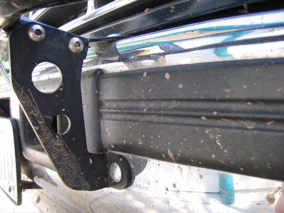 Close-up of a black metal bracket bolted to a vehicle's front bumper. Mud splatters are visible on the bumper and bracket