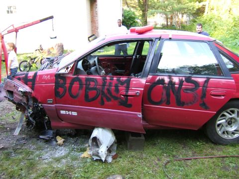 1998 Ford Taurus - A red sedan with extensive graffiti and damage sits in a yard, supported by cinder blocks.