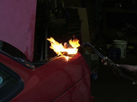 1998 Ford Taurus - A person uses a torch to heat a red car fender in a dimly lit garage.