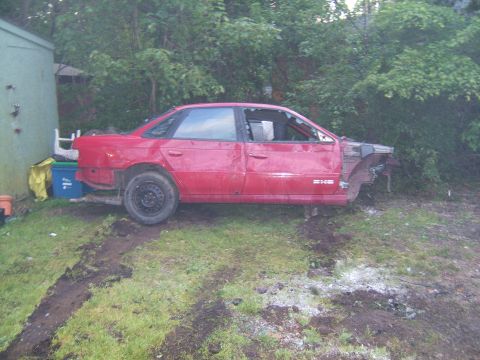 1998 Ford Taurus - This red 1998 Ford Taurus sedan is missing its front end and rear bumper, sitting in a grassy area next to a shed.