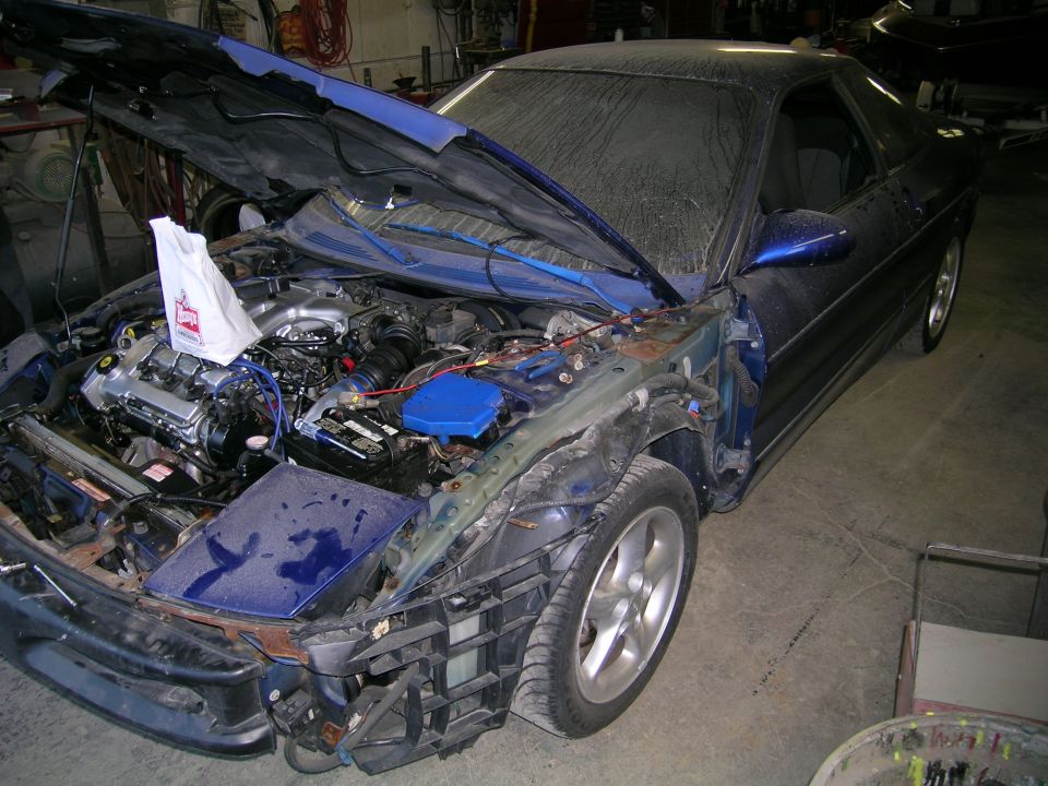 Blue coupe with hood open, showing engine and front-end damage. Dusty windshield and body. Visible Supermotors logo