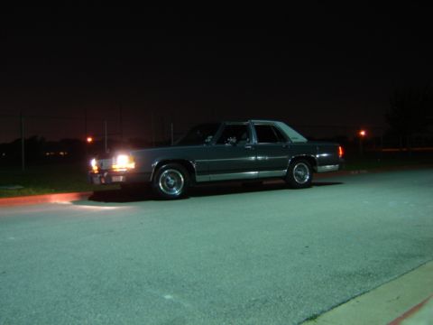 1988 Ford Crown Victoria LTD - A dark sedan with a white roof is parked at night, its headlights illuminating the asphalt.