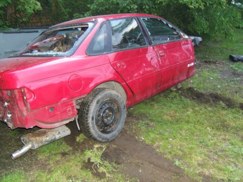 1998 Ford Taurus - Damaged 1998 Ford Taurus sedan sits in a grassy area with broken windows and missing body panels.